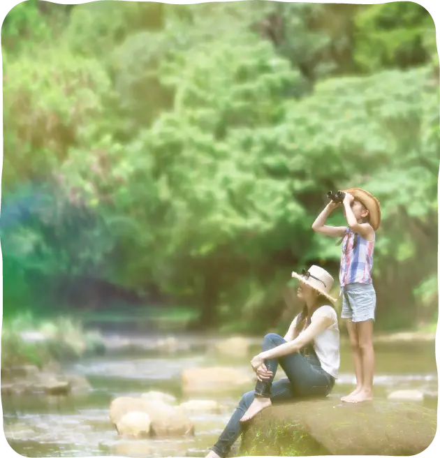 川辺の大きな岩に腰掛けた母親とその隣で双眼鏡で森を眺める女の子